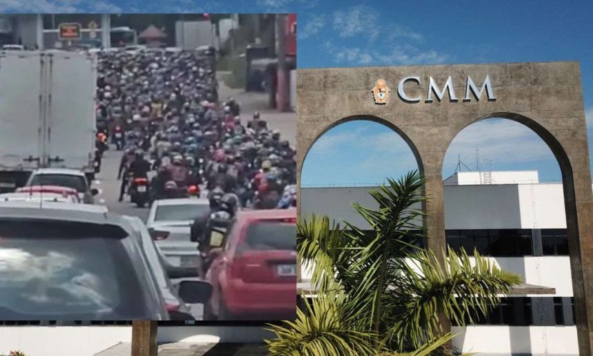 Engarrafamento com motos e carros em frente à CMM, com céu azul e vegetação.