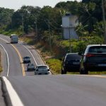 Estrada AM-010 com carros em dia ensolarado, cercada por vegetação densa.
