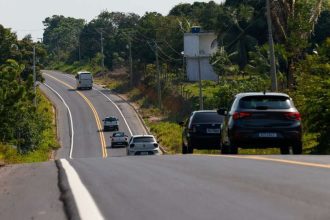 Estrada AM-010 com carros em dia ensolarado, cercada por vegetação densa.