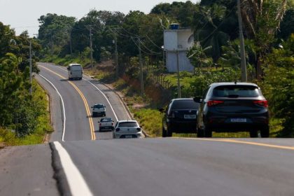 Estrada AM-010 com carros em dia ensolarado, cercada por vegetação densa.