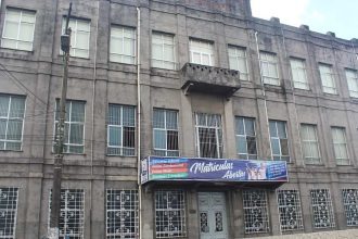 Fachada de escola cinza com letreiro de matrículas abertas.