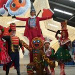 Grupo de artistas fantasiados em cenário natalino, com destaque para a decoração e iluminação.