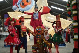 Grupo de artistas fantasiados em cenário natalino, com destaque para a decoração e iluminação.