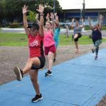 Grupo de mulheres se exercitando em um parque, com os braços erguidos e pernas levantadas.