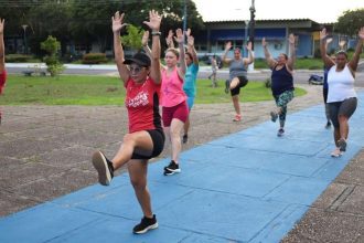 Grupo de mulheres se exercitando em um parque, com os braços erguidos e pernas levantadas.
