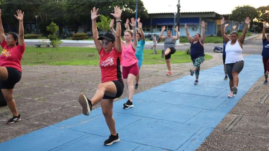 Grupo de mulheres se exercitando em um parque, com os braços erguidos e pernas levantadas.