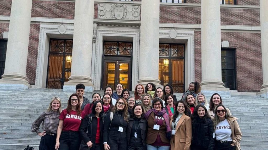 Grupo de pessoas posando em frente a um prédio com colunas, muitas delas vestindo camisas da Harvard.