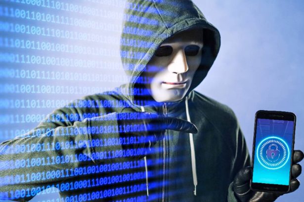 Hacker com máscara e capuz segurando smartphone com cadeado digital, em um fundo de código binário.