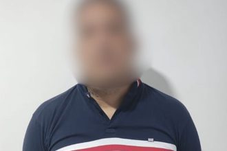 Homem com camisa azul e detalhes em branco e vermelho, rosto desfocado.