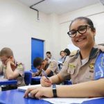Aluna da Polícia Militar sorrindo em sala de aula, com colegas ao fundo.