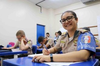 Aluna da Polícia Militar sorrindo em sala de aula, com colegas ao fundo.