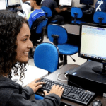 Alunos em aula de informática, com foco em uma jovem sorrindo usando o computador.