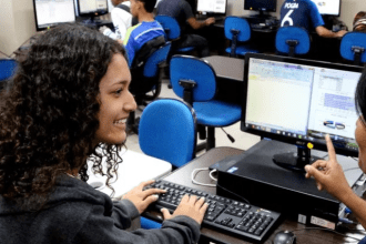 Alunos em aula de informática, com foco em uma jovem sorrindo usando o computador.
