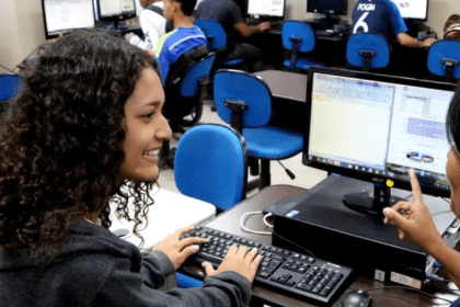 Alunos em aula de informática, com foco em uma jovem sorrindo usando o computador.