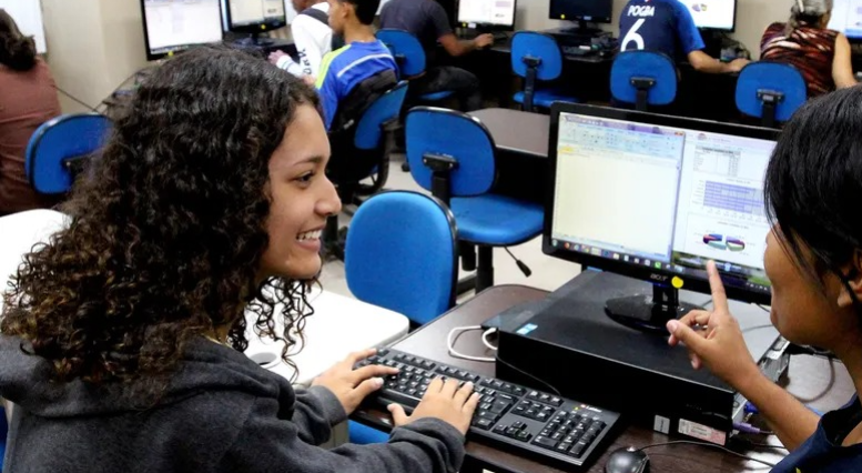 Alunos em aula de informática, com foco em uma jovem sorrindo usando o computador.