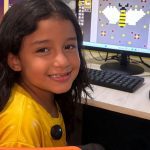 Menina sorrindo em curso de criação de games com um computador mostrando um desenho de abelha em pixels.