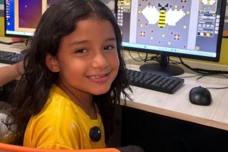 Menina sorrindo em curso de criação de games com um computador mostrando um desenho de abelha em pixels.