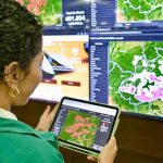 Mulher analisa dados de focos de calor em tablet, com mapas e painéis de dados ao fundo.