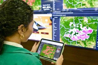 Mulher analisa dados de focos de calor em tablet, com mapas e painéis de dados ao fundo.