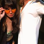 Mulher com óculos laranja e tatuagem de microfone posando ao lado de uma pessoa com camisa branca.