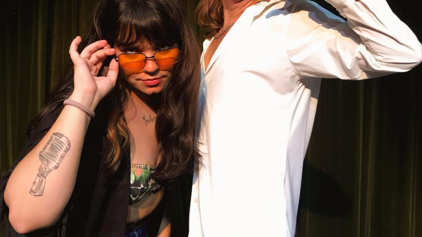 Mulher com óculos laranja e tatuagem de microfone posando ao lado de uma pessoa com camisa branca.