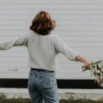 Mulher solteira de costas com flores, em frente a uma parede branca.