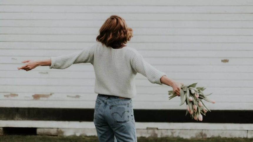 Mulher solteira de costas com flores, em frente a uma parede branca.