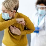 Mulher tossindo em consultório médico, usando máscara e sendo observada por profissional de saúde.