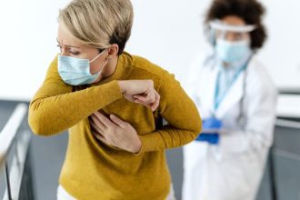 Mulher tossindo em consultório médico, usando máscara e sendo observada por profissional de saúde.