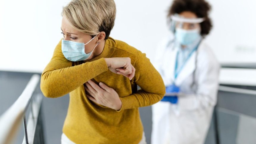 Mulher tossindo em consultório médico, usando máscara e sendo observada por profissional de saúde.