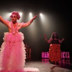 Performance drag no palco do festival, com figurinos elaborados e iluminação dramática.
