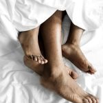 Pés de pessoas na cama, sob lençóis brancos