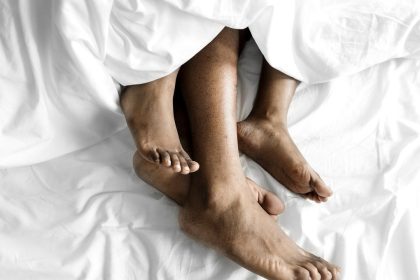 Pés de pessoas na cama, sob lençóis brancos