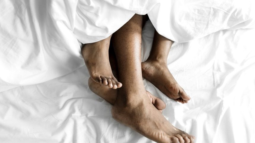 Pés de pessoas na cama, sob lençóis brancos