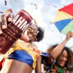 Pessoa segurando lata de cerveja Brahma Duplo Malte em celebração festiva com pessoas e guarda-chuva colorido.