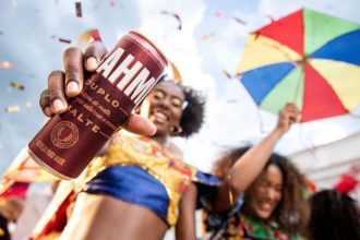 Pessoa segurando lata de cerveja Brahma Duplo Malte em celebração festiva com pessoas e guarda-chuva colorido.
