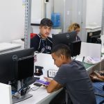 Pessoas trabalhando em escritório com computadores, possivelmente em busca de vagas de emprego.