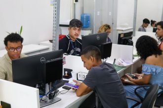 Pessoas trabalhando em escritório com computadores, possivelmente em busca de vagas de emprego.