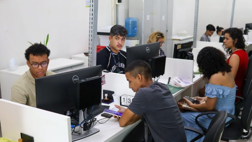 Pessoas trabalhando em escritório com computadores, possivelmente em busca de vagas de emprego.