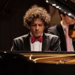 Pianista tocando piano com orquestra ao fundo. Ele está usando um smoking e gravata borboleta.