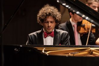 Pianista tocando piano com orquestra ao fundo. Ele está usando um smoking e gravata borboleta.