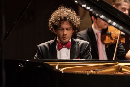 Pianista tocando piano com orquestra ao fundo. Ele está usando um smoking e gravata borboleta.
