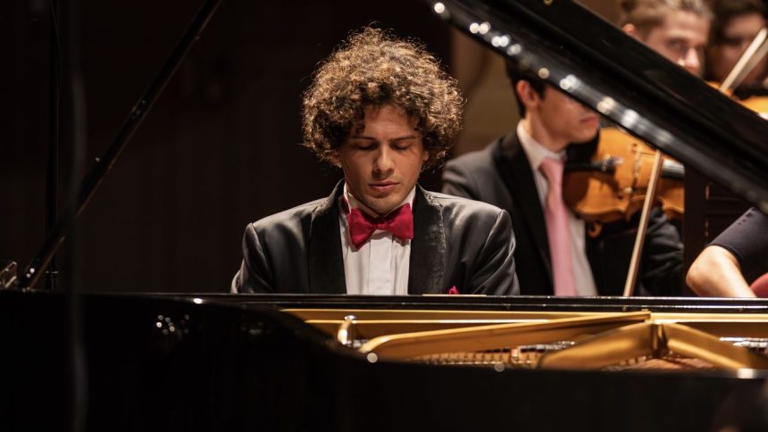 Pianista tocando piano com orquestra ao fundo. Ele está usando um smoking e gravata borboleta.