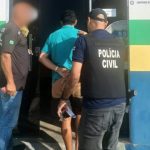 Policial conduz homem preso. O preso está algemado e sendo levado para dentro de um prédio.