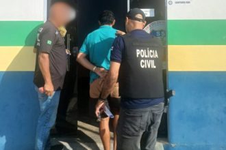 Policial conduz homem preso. O preso está algemado e sendo levado para dentro de um prédio.