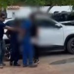 Policial conduz pessoas perto de um carro branco em investigação.