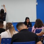 Professora escrevendo em uma lousa, com alunos em sala de aula.