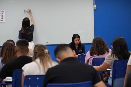Professora escrevendo em uma lousa, com alunos em sala de aula.