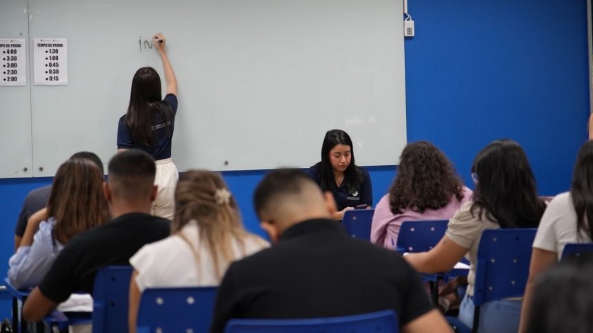Professora escrevendo em uma lousa, com alunos em sala de aula.