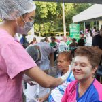 Profissional de saúde aplicando tratamento facial em mulher durante o Arena Rosa Azul, com outras pessoas ao fundo.